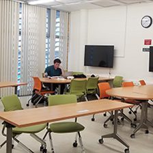 A man sitting alone in a modern classroom with green chairs and a wall-mounted TV.